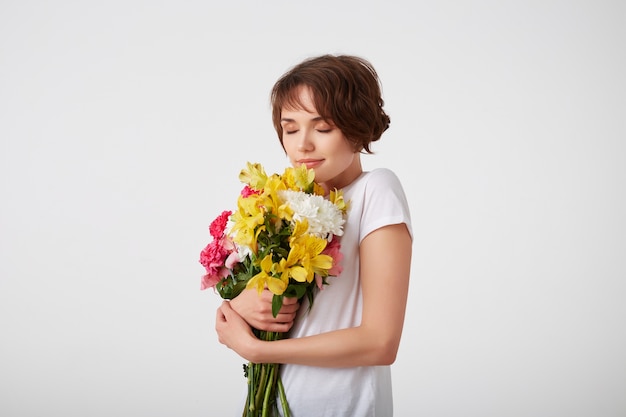 白い空白のtシャツを着て 色とりどりの花の花束を持って においを楽しんで 目を閉じて白い背景の上に立って 若い笑顔の素敵な短い髪の少女 無料の写真