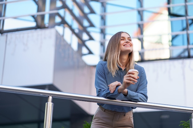 Young smiling professional woman having a coffee break during her full working day. she holds a paper cup outdoors near the business building while relaxing and enjoying her beverage. Free Photo