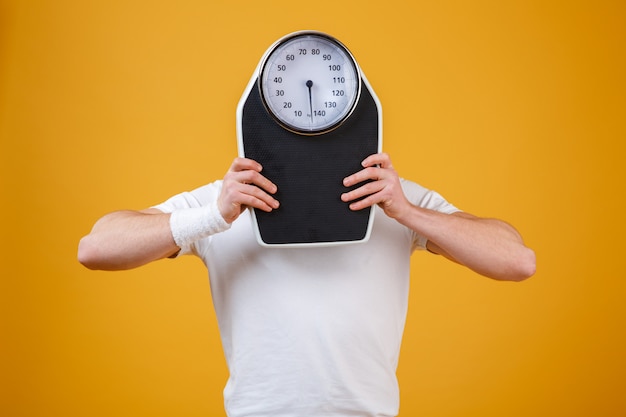 Young sports man hiding face behind weight scales Free Photo