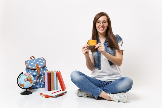 Young surprised joyful woman student in glasses denim clothes holding credit card sitting near globe backpack, school books isolated Free Photo