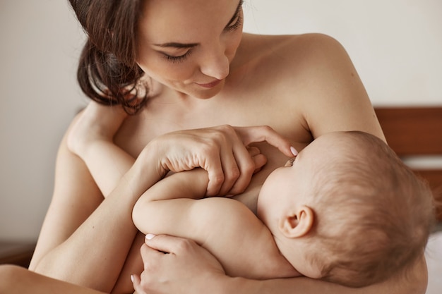 Young tender nude mother breastfeeding hugging her newborn baby sitting in bed at morning Free Photo