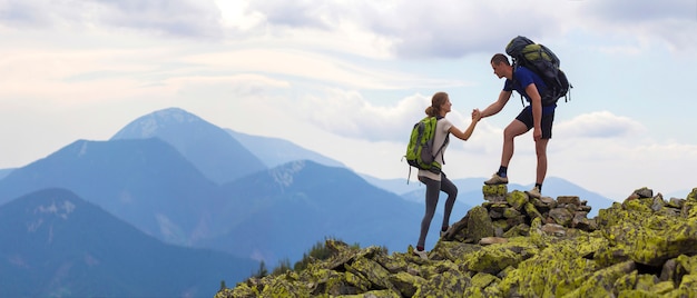 Young tourists with backpacks, athletic boy helps slim girl to clime rocky mountain top against bright summer sky and mountain range background. tourism, traveling and healthy lifestyle concept. Premium Photo