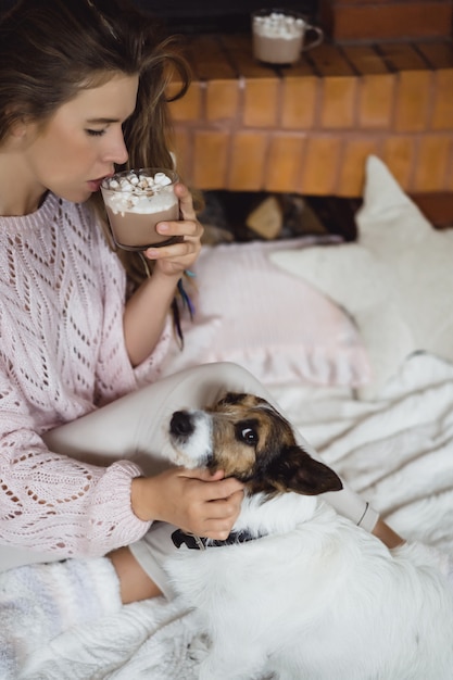 暖炉のそばで若い女性は 犬とマシュメロでココアを飲みます 無料の写真