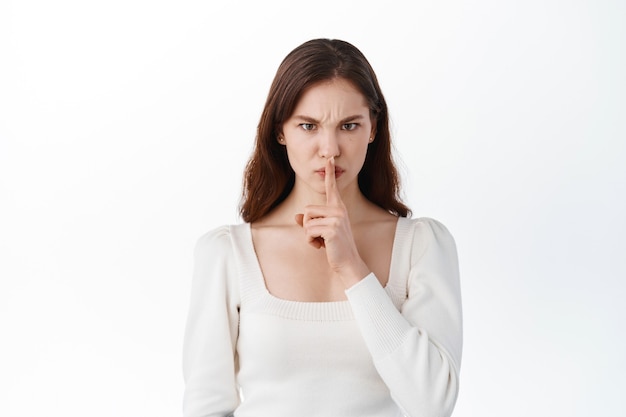 Young woman holding finger on lips mouth to keep it quiet hush, standing isolated on white blank studio wall with copy space, millennial gossip girl showing shh gesture secret in silence Free Photo