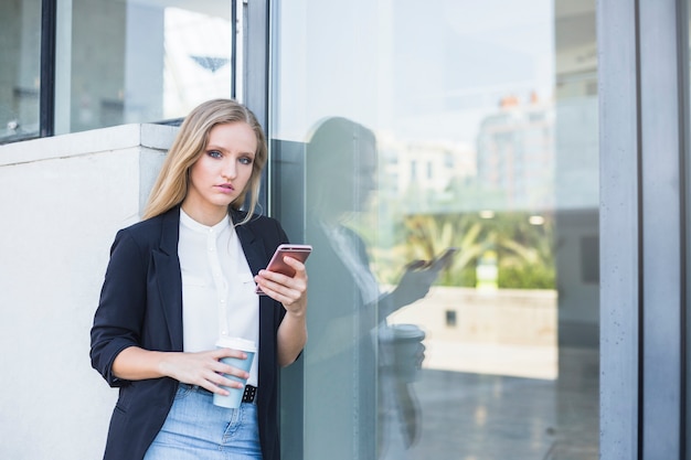 Young woman holding takeaway coffee cup and mobile phone leaning on reflective glass Free Photo