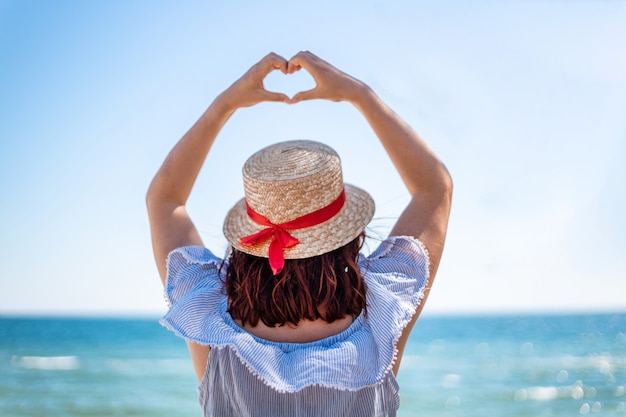 麦わら帽子と夏のドレスの後ろ姿の若い女性は青い海に彼女の手で愛のサインを作る プレミアム写真
