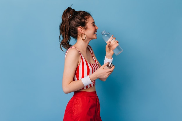Young woman in red striped outfit laughs and drinks water from bottle on isolated wall Free Photo