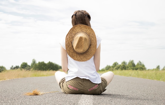 麦わら帽子をかぶった 荒野の無限のまっすぐな空の道に座っている若い女性 プレミアム写真