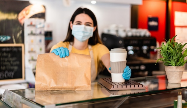 Young woman wearing face mask while serving takeaway breakfast and coffee inside cafeteria restaurant Premium Photo