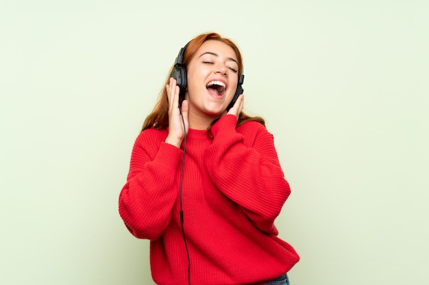 Adolescente rousse avec pull sur vert isolé, écouter de la musique avec des écouteurs Photo Premium