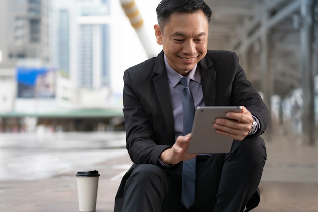Asiatique homme d'affaires assis et tenant une tablette numérique avec des immeubles de bureaux dans la ville Photo Premium