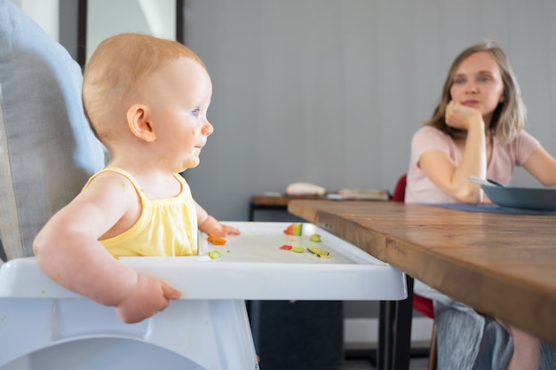 Beau Nouveau Ne Rousse Mangeant Et Assis Dans Un Siege De Cuisine Pour Bebe En Plastique Blanc Belle Jeune Mere Assise A Une Table En Bois Et Regardant Son Enfant Concept De Famille