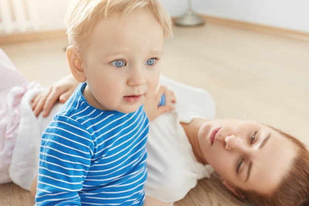 Beau Portrait De Petit Garcon Blond Aux Yeux Bleus Et Mere Attentionnee Allonge Sur Le Sol A La Maison Petit Bebe En Vetements Bleus Regardant Vers L Avant Sa Jolie Maman Attentive Le