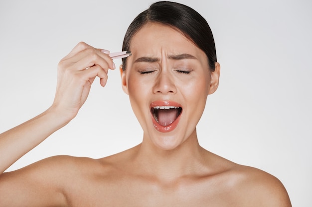 Beauté De La Jeune Femme Stressée Aux Cheveux Bruns Hurlant De Douleur Tout En Arrachant Les Sourcils à L'aide De Pinces, Isolé Sur Blanc Photo gratuit