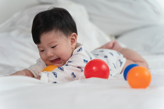 Bebe Asiatique Rit Et Joue A La Balle Avec Des Jouets Sur Un Lit Blanc Avec Un Sentiment De Joie Et De Gaiete Et Le Bebe Qui Rampe Sur Le Lit