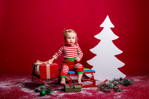 Bebe En Pyjama De Noel Et Bonnet De Noel Attrape La Neige Assis Sur Un Traineau Avec Boite Cadeau Et Grand Arbre De Noel Blanc Sur Fond Rouge Photo Premium