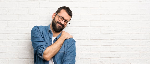Bel homme à la barbe sur mur de briques blanches souffrant de douleur à l'épaule pour avoir fait un effort Photo Premium