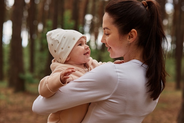 Belle Femme Aux Cheveux Noirs Portant Des Vetements Blancs Posant En Plein Air Tenant Un Bebe Dans Les Mains Et Regardant Sa Fille Avec Beaucoup D Amour Jouant Dans La Foret Photo