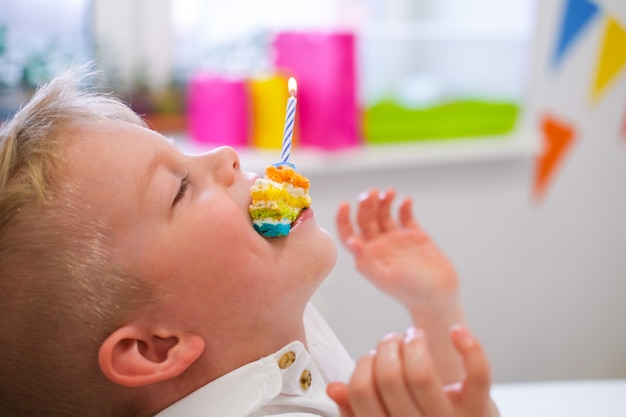 Blond Caucasien Garcon S Amuse Et Tient Un Morceau De Gateau Arc En Ciel D Anniversaire Avec Une Bougie Allumee Dans Sa Bouche A La Fete D Anniversaire Fond Colore Festif Photo Premium