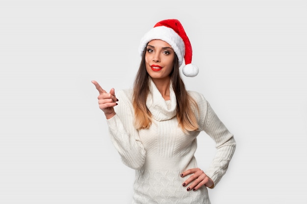 Bouchent Portrait Belle Femme Caucasienne En Bonnet Rouge Sur Mur Blanc. Concept De Noël Nouvel An. Mignon Femme Dents Souriant Positif Montrant L'index Du Côté Du Doigt Photo Premium