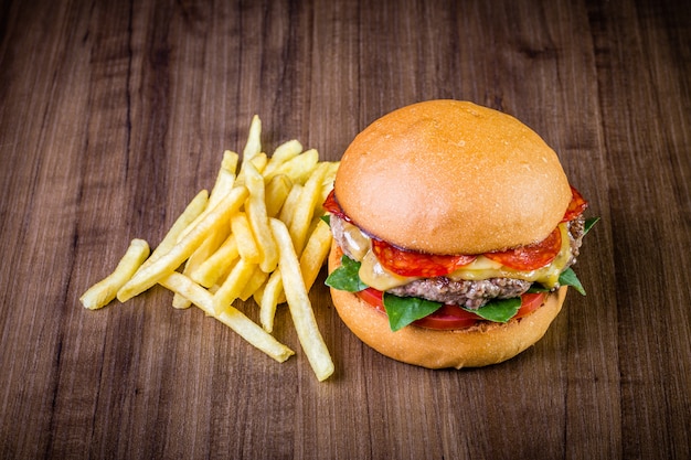 Burger de bœuf au fromage, peperoni italien, tomate, feuilles de basilic et frites sur table en bois Photo Premium