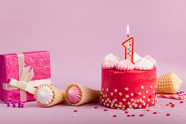 Chandelle allumée numéro un sur un délicieux gâteau rouge avec des étoiles saupoudrées; cônes de gaufres et boîte-cadeau sur fond violet Photo gratuit