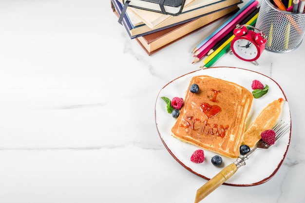 Concept de petit-déjeuner pour la rentrée des classes, crêpes à la confiture de framboises - j'aime l'école, une étole en marbre blanc, des livres, un réveil, des crayons, des fournitures scolaires. vue de dessus Photo Premium
