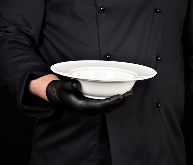 Cuisinier en uniforme noir et gants en latex noir tient dans sa main une assiette blanche vide ronde Photo Premium