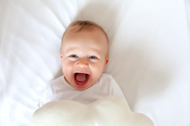 Drole Blond Mignon Petit Bebe Aux Yeux Bleus En Body Blanc Couche Et Riant Dans Le Lit Des Enfants Blancs Photo Premium