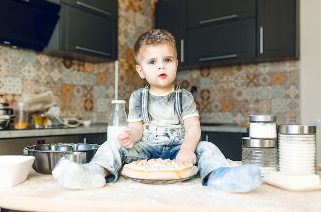 Drole Enfant Assis Sur La Table De La Cuisine Dans Une Cuisine Roustique Jouant Avec De La Farine Et Degustant Un Gateau Photo Gratuite