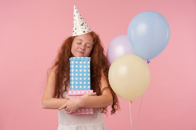 Enfant De Femme Frisee Rousse Positive Posant Sur Fond De Studio Rose Avec Des Ballons Colores Tenant Des Coffrets Cadeaux Avec Les Yeux Fermes Et Souriant Joyeusement Portant Des Vetements De Fete