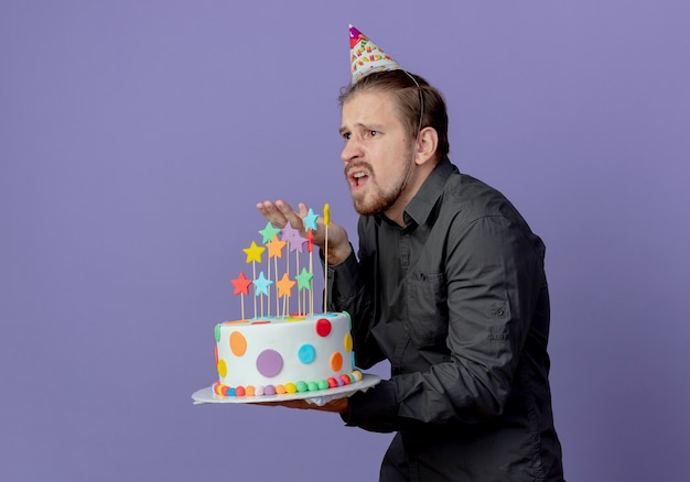 Ennuye Bel Homme Dans Le Chapeau D Anniversaire Se Tient Sur Le Cote Tient Le Gateau Pointant Vers L Avant Isole Sur Mur Violet Photo Gratuite