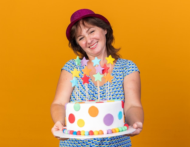 Femme D Age Moyen En Chapeau De Fete Tenant Un Gateau D Anniversaire Avec Le Sourire Sur Le Visage Heureux Et Joyeux Celebrant La Fete D Anniversaire Debout Sur Le Mur Orange Photo Gratuite