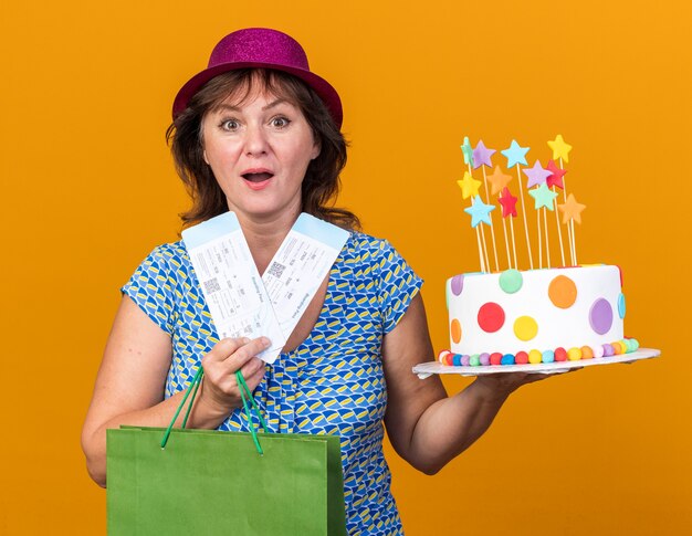 Femme D Age Moyen En Chapeau De Fete Tenant Un Sac En Papier Avec Des Cadeaux Tenant Un Gateau D Anniversaire Et Des Billets D Avion Heureux Et Surpris Photo Gratuite