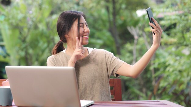 Femme asiatique utilisant un téléphone portable post selfie dans les médias sociaux, femme relax se sentir heureux montrant des sacs à provisions assis sur une table dans le jardin le matin. Photo gratuit