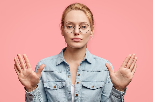 Femme blonde sérieuse avec une expression confiante, montre des paumes, fait un geste d'arrêt, ne permet pas de faire quelque chose, porte des vêtements à la mode, isolés sur un mur rose Photo gratuit