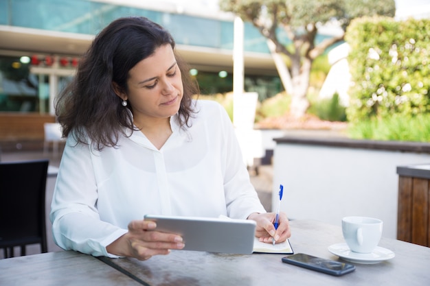 Femme ciblée, prendre des notes, travailler et utiliser une tablette au café Photo gratuit