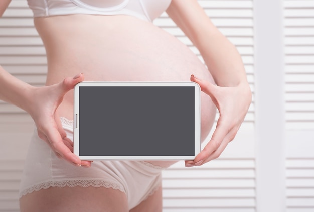 Femme enceinte en tenue tenant et montrant une tablette numérique avec affichage vide Photo Premium