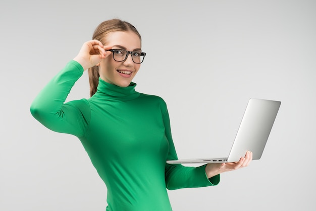 Femme joyeuse à lunettes travaille sur un ordinateur portable en le tenant dans ses mains tout en se tenant contre le fond blanc. regarde la caméra Photo Premium