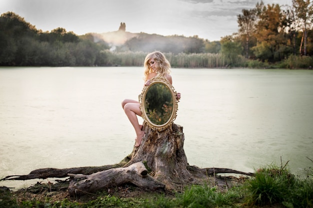 Femme nue blonde tient un miroir assis devant un lac couvert de brume Télécharger des Photos