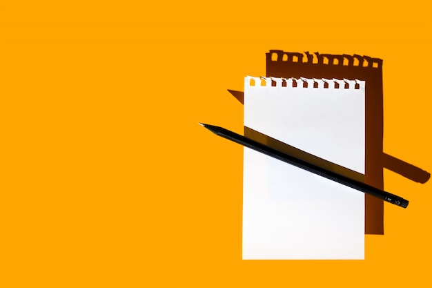 Une feuille vierge de bloc-notes, un crayon noir et des ombres dures sur jaune vif Photo Premium