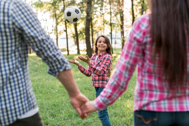 Fille jouant avec un ballon de soccer devant les parents se tenant la main dans le parc Photo gratuit