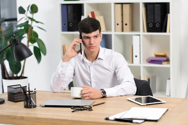 Gros plan, de, sérieux, jeune homme affaires, tenue, tasse café, parler, sur, téléphone portable, sur, lieu de travail Photo gratuit
