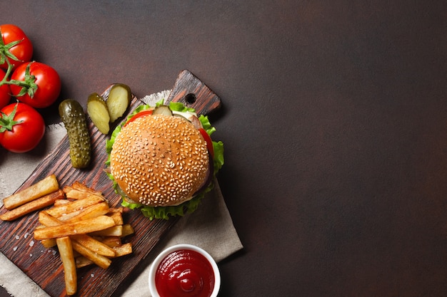 Hamburger fait maison avec du bœuf, des tomates, de la laitue, du fromage, des oignons, des concombres et des frites sur une planche à découper et un fond rouillé Photo Premium
