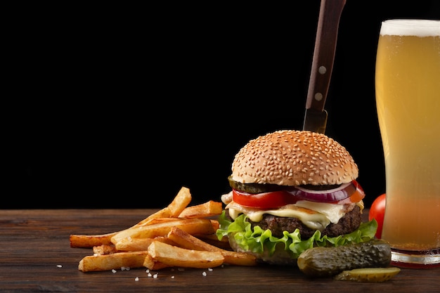 Hamburger fait maison avec des frites et un verre de bière sur une table en bois. dans le burger coincé un couteau. Photo Premium