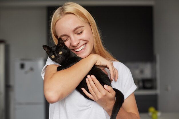 Heureuse Jeune Femme Blonde A La Cuisine Tient Sur Les Mains Caresses Caresses Calins Petit Chat Noir Mignon Souriant Tendrement Photo Gratuite