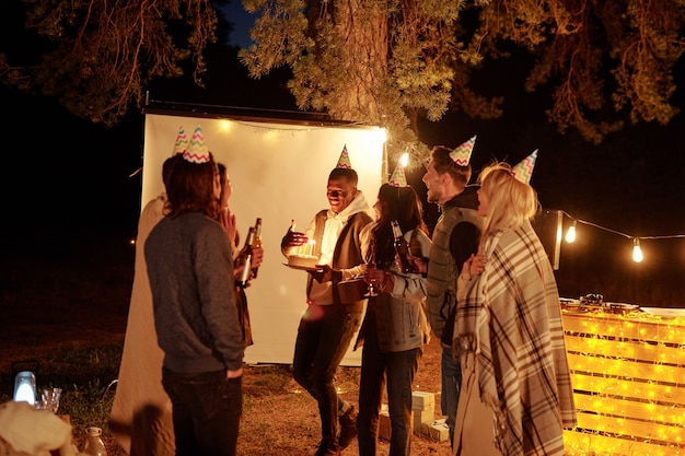 Heureux Jeune Homme Africain Gardant La Main Par Des Bougies Sur Le Gateau D Anniversaire En Se Tenant Debout Entre Amis En Casquettes De Grillage Avec Des Bouteilles De Biere Photo Premium