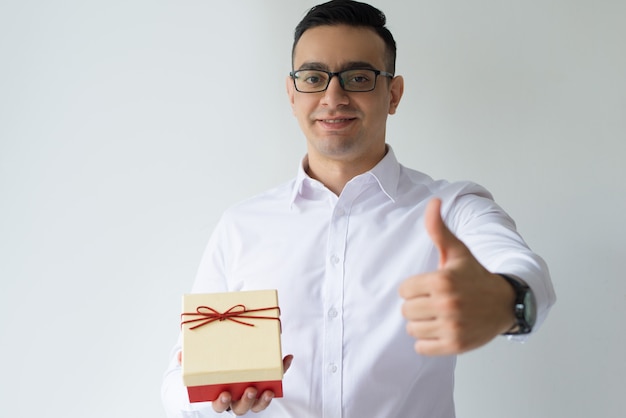 Homme d'affaires souriant tenant une boîte-cadeau et montrant le pouce vers le haut Photo gratuit