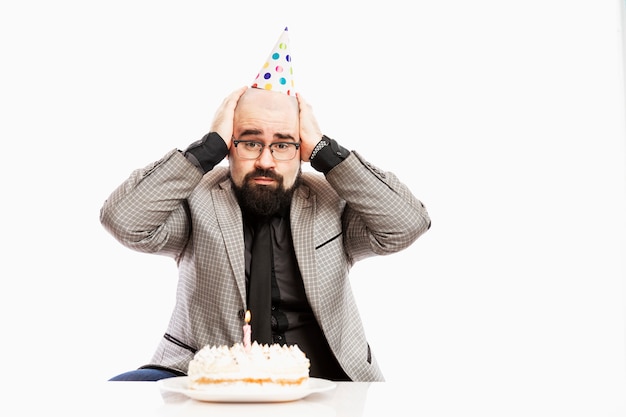 Un Homme Au Chapeau De Clown Est Assis Devant Un Gateau Avec Une Bougie Triste Anniversaire Mur Blanc Photo Premium