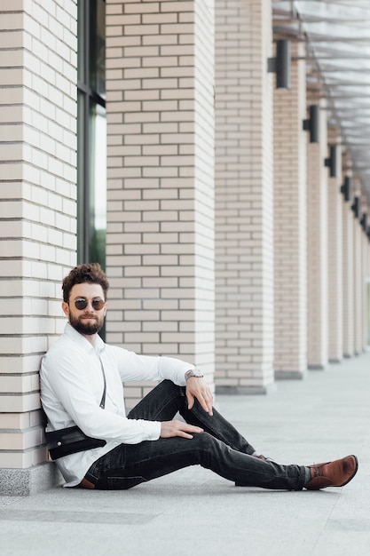 Un Homme Barbu Heureux Souriant Et Elegant Assis Sur La Farine En Chemise Blanche Dans Les Rues De La Ville Pres Du Centre De Bureaux Moderne Photo Gratuite